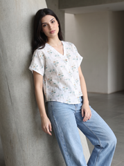 A woman wearing a white short-sleeve V-neck top with a delicate blue and green floral pattern, paired with light-wash blue jeans, leaning against a concrete pillar in a minimalist indoor setting.