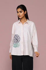 A woman modeling a light pink long-sleeve button-up shirt with a black and green tropical leaf graphic on the front, paired with black pants, against a solid dusty pink background.