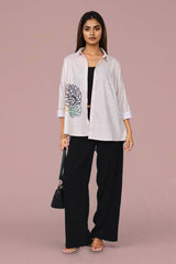 A woman models a white long-sleeve button-up shirt with a large black and green floral graphic on the left sleeve, paired with black wide-leg pants and a small black handbag, set against a solid dusty pink studio background.