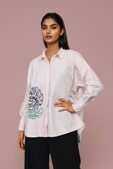 A woman modeling a light pink button-up shirt with a large black and green tropical leaf graphic on the front pocket, paired with black pants, against a solid dusty pink background.