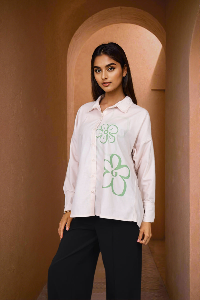 A woman stands in a warm-toned arched corridor wearing a light pink button-up shirt with green floral line drawings and black pants.