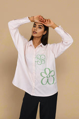 A woman modeling a light pink long-sleeve button-up shirt with green outlined flower graphics on the front, paired with black pants, against a solid beige background.