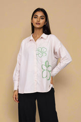 A woman modeling a light pink button-up shirt with green floral line-art graphics on the front, paired with black pants, against a solid beige background.