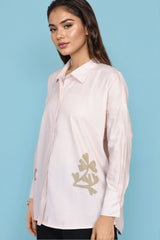 A woman wearing a light pink long-sleeve button-up shirt with gold floral embroidery on the lower front and sleeve, posed against a solid light blue background.