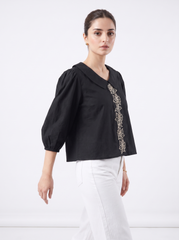 A woman modeling a black cropped blouse with voluminous puff sleeves and a vertical embroidered detail in beige and gold along the front, paired with white trousers, against a plain white studio background.