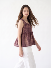 A woman modeling a sleeveless tiered peplum top with a brown and white gingham check pattern, paired with white wide-leg pants, against a plain white studio background.