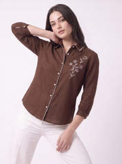 A woman modeling a brown long-sleeve button-up shirt with white floral embroidery on the chest and white piping along the placket, paired with white pants against a light studio background.