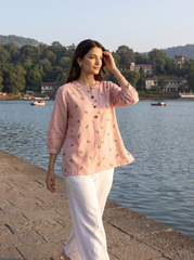 A woman wearing a light pink embroidered kurta and white trousers stands on a stone walkway next to a lake, with boats and a tree-lined shore in the background.