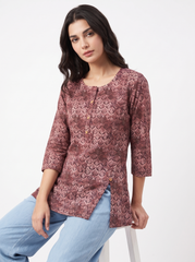 A woman wearing a dusty rose and brown patterned kurta with three-quarter sleeves, paired with light blue jeans, seated against a plain white studio background.