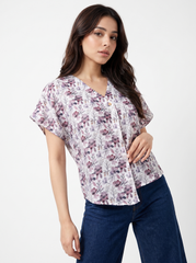 A woman modeling a short-sleeve V-neck blouse with a muted floral print in dusty purple, mauve, and gray on a light background, paired with dark blue jeans against a plain light gray studio background.