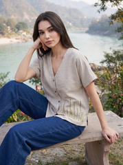 A woman sits on a wooden bench outdoors by a river, wearing a light beige short-sleeve button-front top with a small embroidered floral detail near the hem and dark blue jeans.