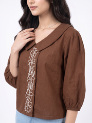 A woman wearing a brown wrap-style blouse with white floral embroidery down the front, paired with light blue jeans against a plain white background.