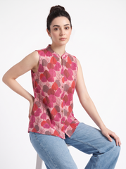 A woman wearing a sleeveless pink and red patterned top with a stand collar, paired with light blue jeans, posing against a plain white studio background.