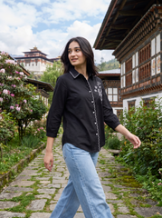 A woman wearing a black long-sleeve button-up shirt with white embroidery and light blue jeans, walking along a stone path in a traditional village setting with buildings and greenery in the background.