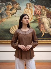 A woman wearing a brown long-sleeve top with a detailed embroidered neckline and white pants, standing in front of a large classical painting.