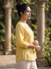 A woman wearing a light yellow linen button-up top with small green dragonfly prints and off-white trousers, standing outdoors in front of stone pillars and green foliage.