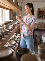 A woman in a pottery studio wearing a floral print short-sleeve shirt and blue jeans, holding a small handmade ceramic bowl. Shelves filled with various ceramic bowls and plates are visible in the background and foreground.