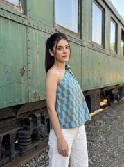 A woman posing in front of an old green train car, wearing a light blue and gray checkered halter top and white pants.