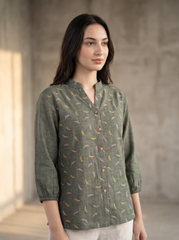 A woman wearing a sage green linen button-up shirt with a delicate multicolor floral pattern, standing in a minimalist concrete room.