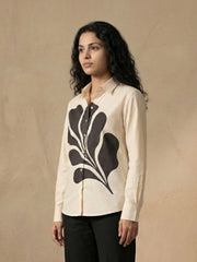 A woman modeling a cream-colored long-sleeve button-up shirt with a large black abstract leaf pattern on the front, paired with dark gray trousers, set against a textured beige background.