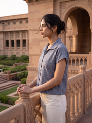 A woman stands in profile on a stone balcony wearing a short-sleeve textured blue button-up shirt and cream trousers, with a large sandstone building and manicured gardens in the background.