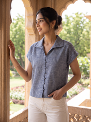 A woman wearing a short-sleeve button-front linen shirt in a textured slate blue-gray, paired with off-white trousers, standing on a stone balcony with a garden background.