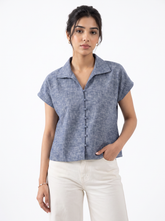 A woman modeling a short-sleeve button-front collared top in a heathered blue-gray fabric, paired with off-white pants, against a plain white studio background.