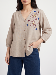 A woman wearing a beige linen button-up blouse with colorful floral embroidery on the chest and dark blue jeans, photographed against a clean white studio background.