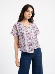 A woman modeling a short-sleeve V-neck blouse with a muted floral print in dusty pink, purple, and beige on a white base, paired with dark blue jeans, against a plain white studio background.