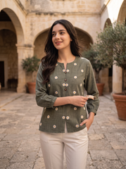 A woman stands in a stone courtyard wearing a sage green long-sleeve top with white polka dots and white pants.