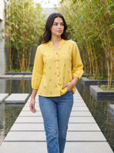 A woman walking on a stone path in a garden setting, wearing a yellow button-up shirt with small heart prints and blue jeans.
