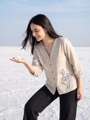 A woman wearing a beige linen button-up shirt with a blue floral embroidery on the lower right side, paired with black pants, standing in a snowy landscape and holding a small white flower in her hand.