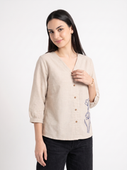 A woman modeling a light beige linen button-up blouse with a V-neck, 3/4 sleeves, and blue floral embroidery on the side, paired with black jeans against a plain white studio background.