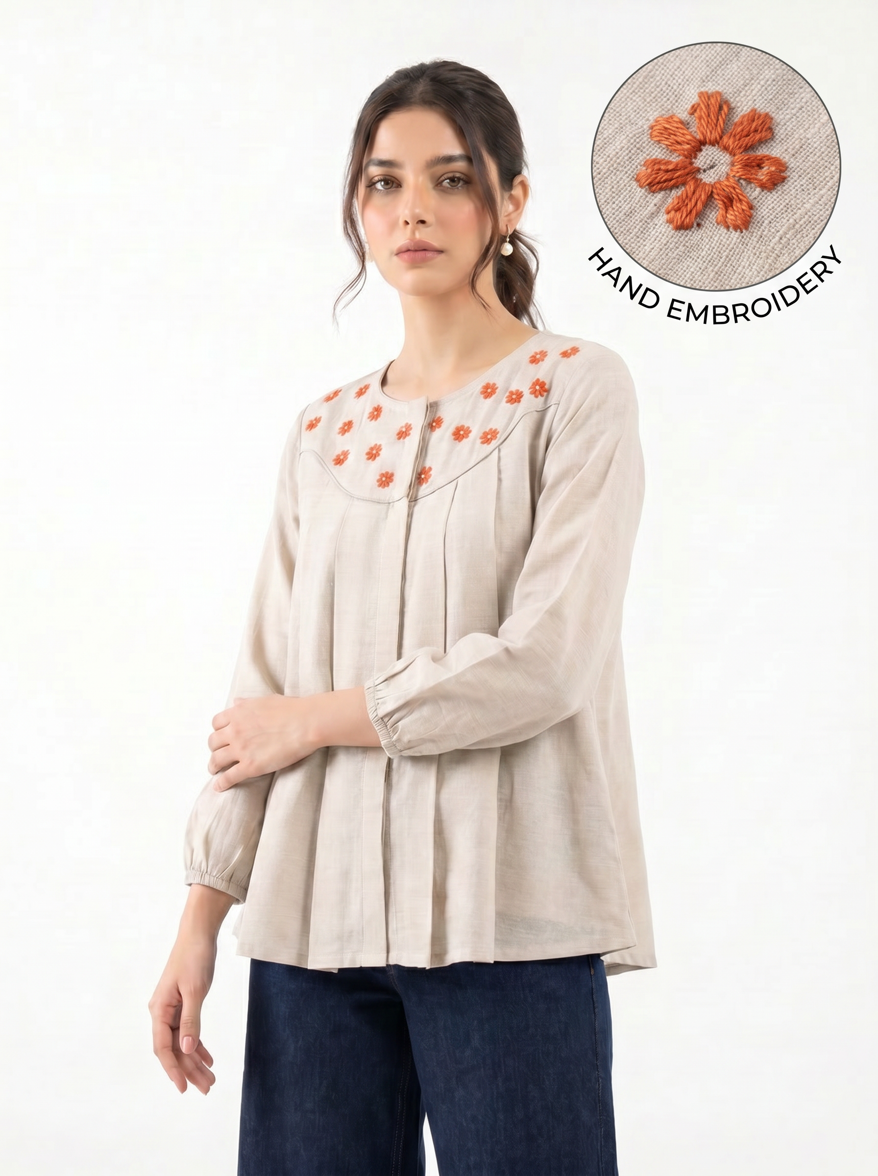 A woman wearing a light beige long-sleeve blouse with orange hand-embroidered details around the neckline, paired with dark blue pants. An inset circle in the top right shows a close-up of the orange embroidery on beige fabric with the text 'HAND EMBROIDERY'.