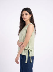 A woman modeling a light sage green sleeveless top with a side tie detail, paired with dark blue jeans, against a plain white studio background.