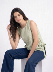 A woman with long dark wavy hair sits on a white block against a white textured wall, wearing a sage green sleeveless top with white embroidered details and dark blue wide-leg jeans.