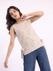 A woman modeling a sleeveless beige linen top with small yellow and pink embroidered flowers and side tie details, paired with blue jeans, against a plain white background.