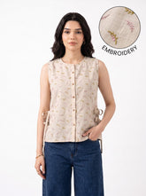 A woman modeling a sleeveless beige top with a subtle embroidered pattern, paired with blue jeans, against a plain white background. An inset circle in the top right highlights the embroidery detail with the text 'EMBROIDERY'.
