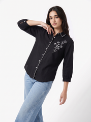 A woman modeling a black long-sleeve button-up shirt with white floral embroidery on the chest and a decorative white chain detail along the front placket, paired with light blue jeans against a plain white studio background.