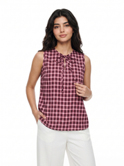 A woman modeling a sleeveless gingham tie-neck blouse in a pink and dark red check pattern, paired with white pants, against a plain white background.