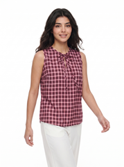 A woman modeling a sleeveless red and black gingham tie-neck blouse with white pants, shot against a solid white background.