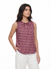 A woman modeling a sleeveless gingham top in a dusty rose and white check pattern with a tie-neck detail, paired with white pants, against a plain white background.