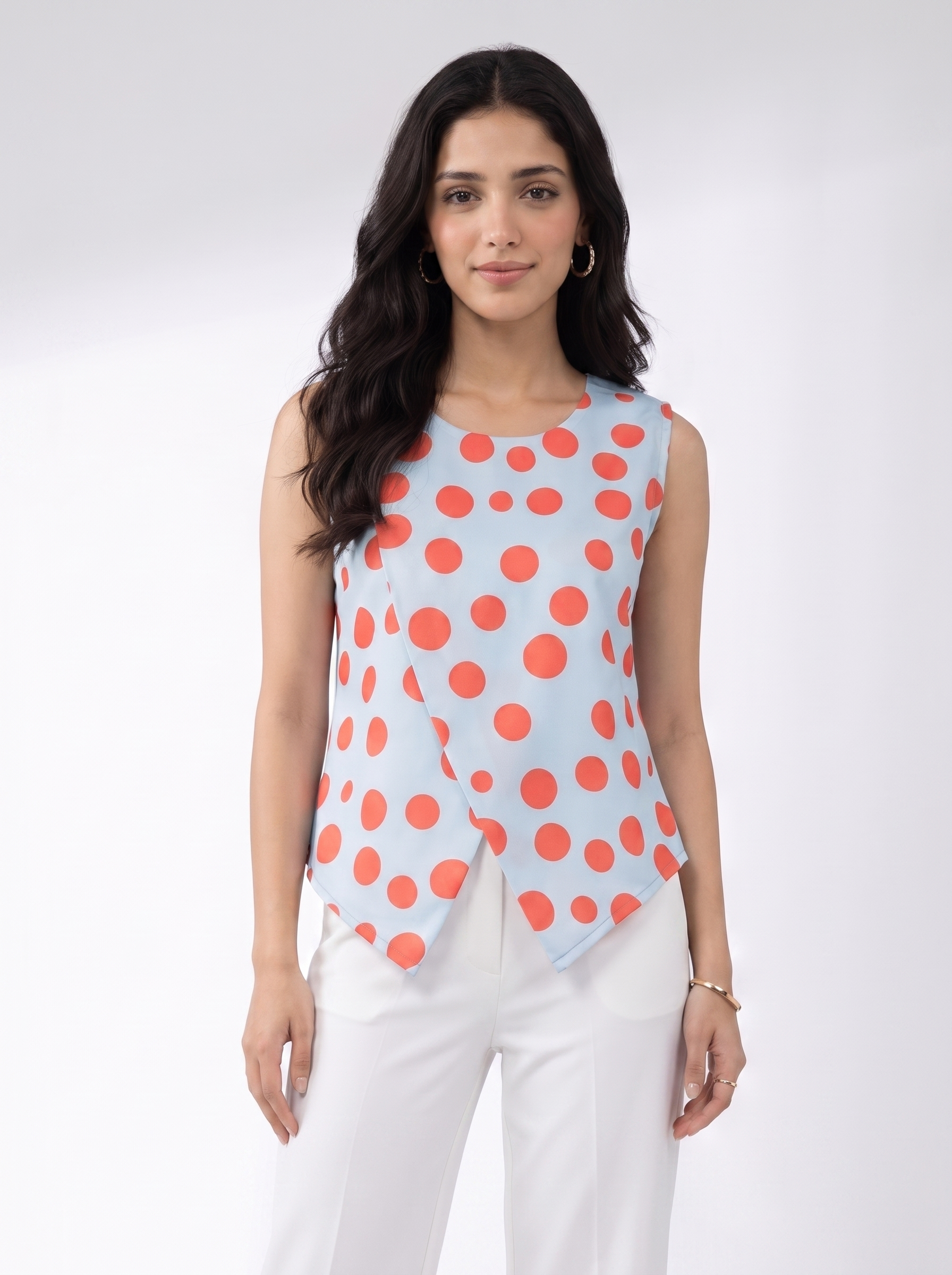 A woman modeling a sleeveless wrap-style top with a light blue base and large coral-red polka dots, paired with white pants, against a plain light gray studio background.