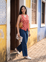 A woman stands on a cobblestone street in front of a colorful yellow and blue building, wearing a red sleeveless top with white polka dots, blue jeans, white loafers, and carrying a tan shoulder bag while holding a magazine.