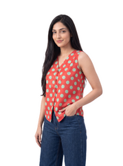 A woman modeling a sleeveless coral-red top with large mint green polka dots, paired with blue jeans, against a solid white background.