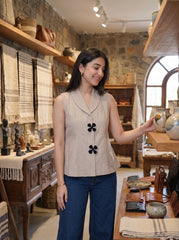 A woman stands inside a rustic shop with stone walls and wooden shelves, wearing a sleeveless beige top with black flower-shaped buttons and dark blue jeans. She is gesturing toward a table display.