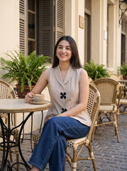 A woman sitting at a wicker cafe table holding a cup of coffee, wearing a beige sleeveless top with black floral details and blue jeans, set against a background of a cafe with shutters and plants.
