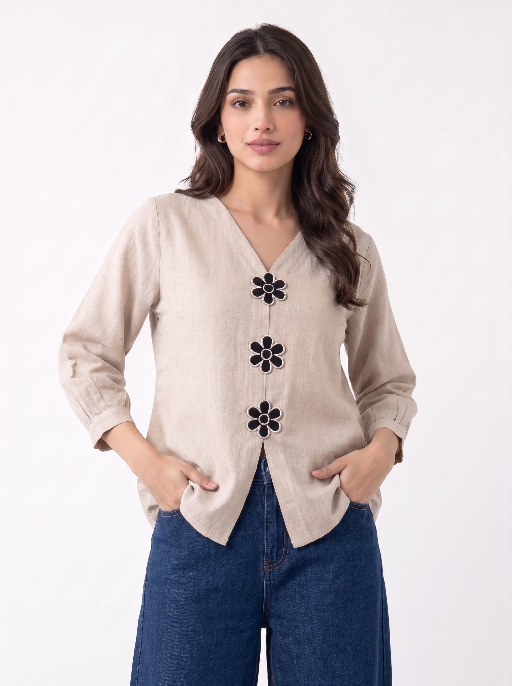 A woman modeling a beige linen-style cardigan with black embroidered flower details down the front, paired with blue jeans, against a plain white background.