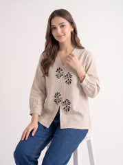 A woman wearing a beige long-sleeve linen top with black floral embroidery and blue jeans, standing in front of a warm, arched stone architectural background.