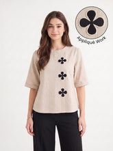 A woman modeling a beige short-sleeve top with three black floral appliques down the front, paired with black pants. A circular inset in the top right shows a close-up of the black-on-beige floral applique detail with the text 'Appliqué Work'.